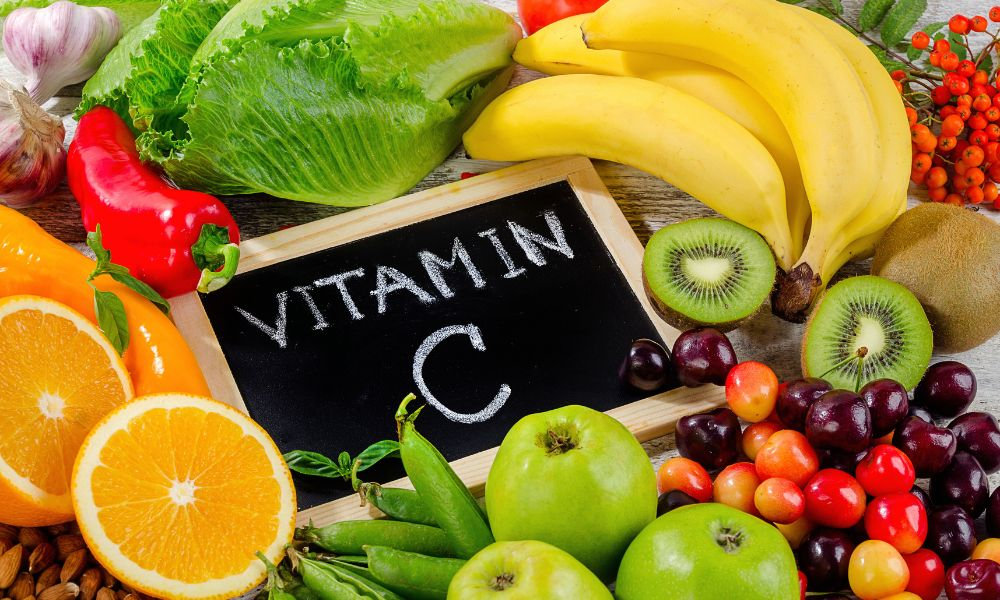  Ovocie a zelenina rozložené okolo nápisu vitamín C znázorňujú význam vitamínu C pre imunitný systém, zdravú pokožku a ochranu buniek pred voľnými radikálmi.