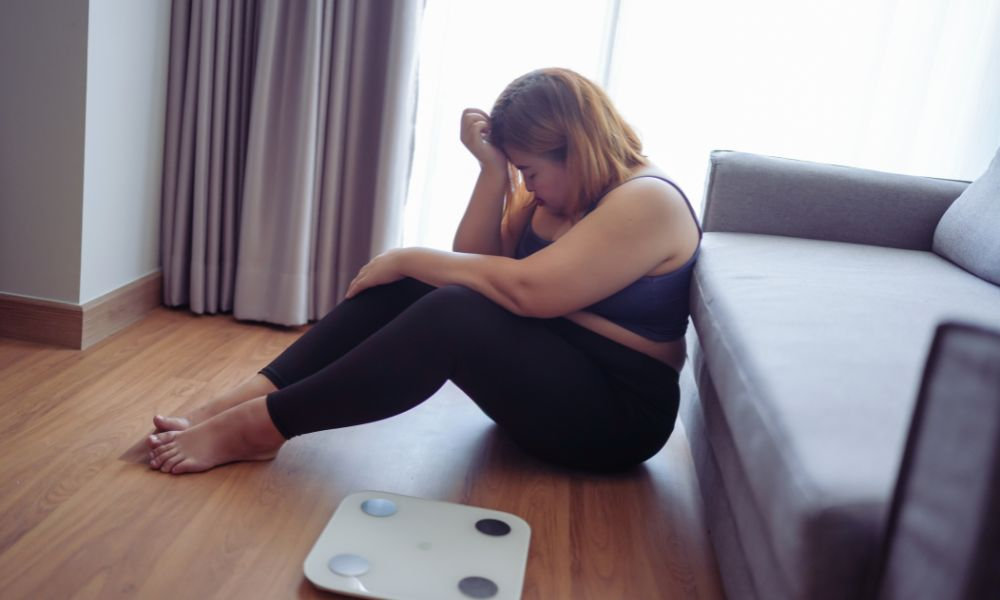  Žena smutná z nadváhy – Sedí na zemi vedľa gauča so sklopenou hlavou a pred sebou má váhu