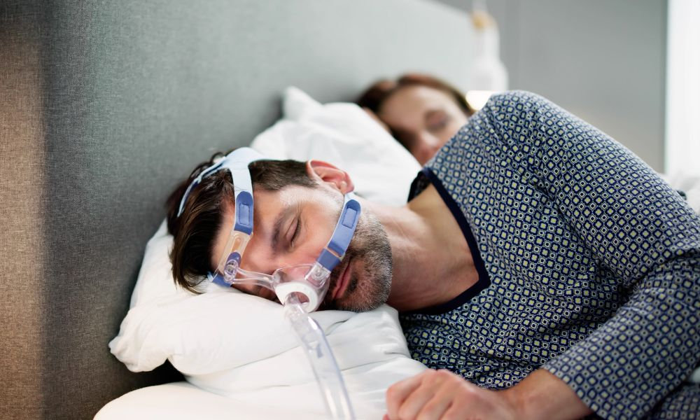 Muž spí v posteli vedľa partnerky s CPAP maskou na tvári pripojenou na hadičku, ktorá patrí medzi najúčinnejšie lieky na apnoe a liečbu spánkového apnoe.