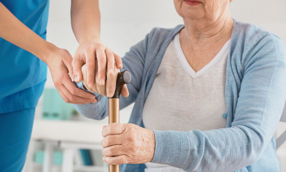  Staršia žena používajúca palicu s asistenciou zdravotníckeho personálu – prevencia a podpora pacientov s Parkinsonovou chorobou.