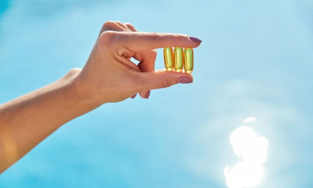  Ruka držiaca kapsuly na pozadí slnečnej oblohy symbolizuje vitamín D získavaný zo slnečného žiarenia a doplnkov výživy, dôležitý pre imunitu, kosti a správnu funkciu organizmu.