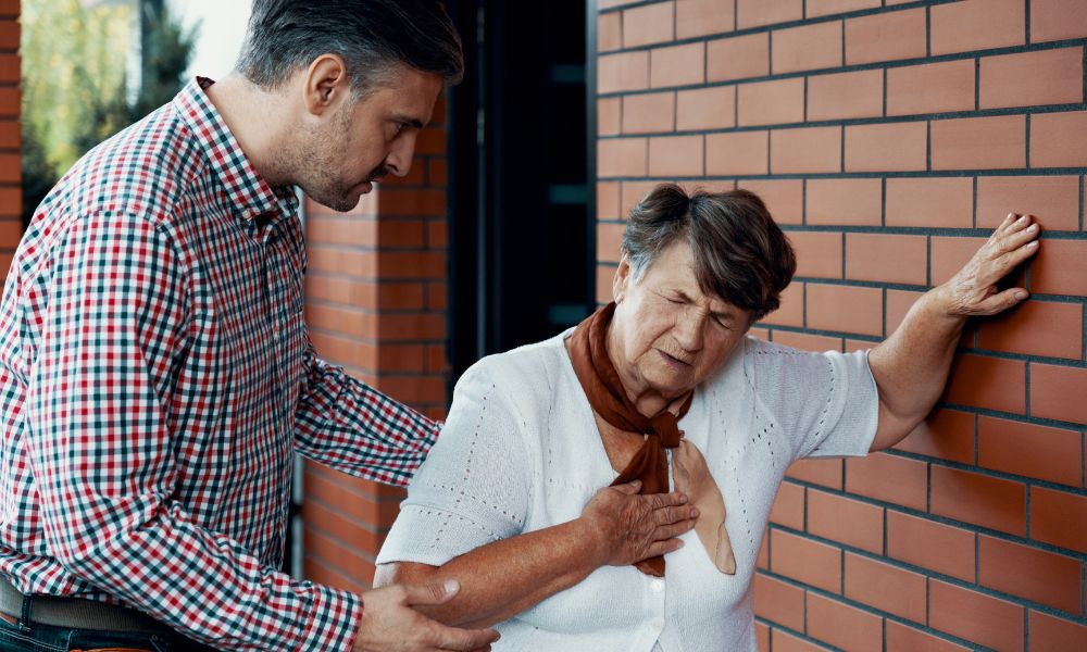  Staršia žena s ťažkosťami pri dýchaní si drží ruku na hrudi a opiera sa o tehlovú stenu, zatiaľ čo jej muž pridržiava, čo zobrazuje akútnu dýchavičnosť ako možný príznak dyspnoe.